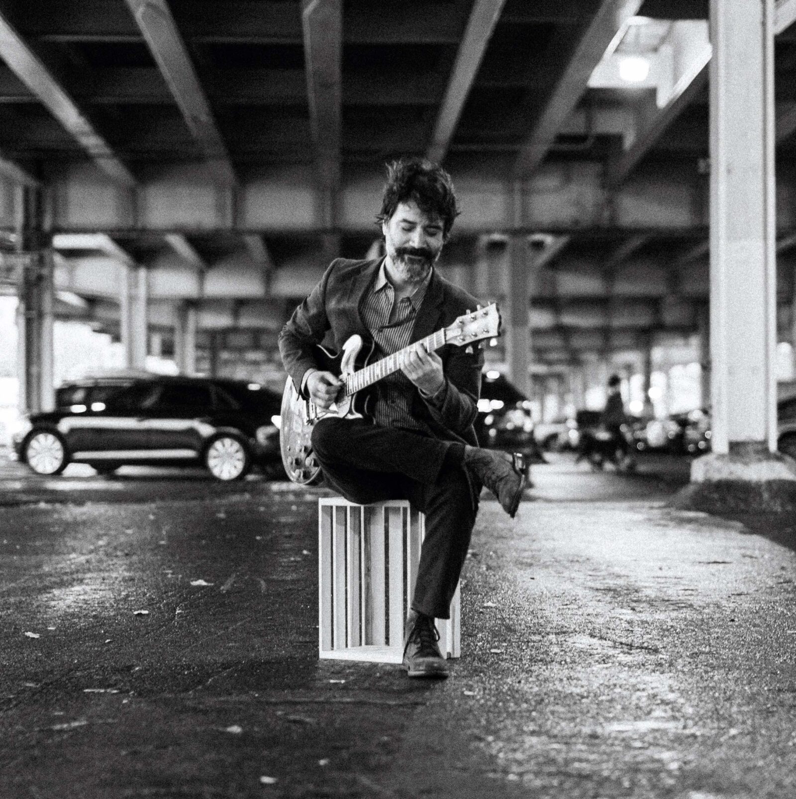 Photo of Gui Monteiro holding sitting and playing his electric guitar in a parking lot