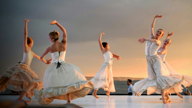 Five dancers in flowing white dresses perform an outdoor contemporary ballet on a white stage at sunset.