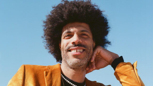 Portrait of jazz trumpeter Wayne Tucker smiling in a mustard blazer against a blue sky.
