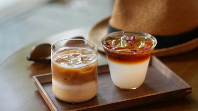 Two iced coffee drinks on a wooden tray, one layered with milk and one topped with rose petals.