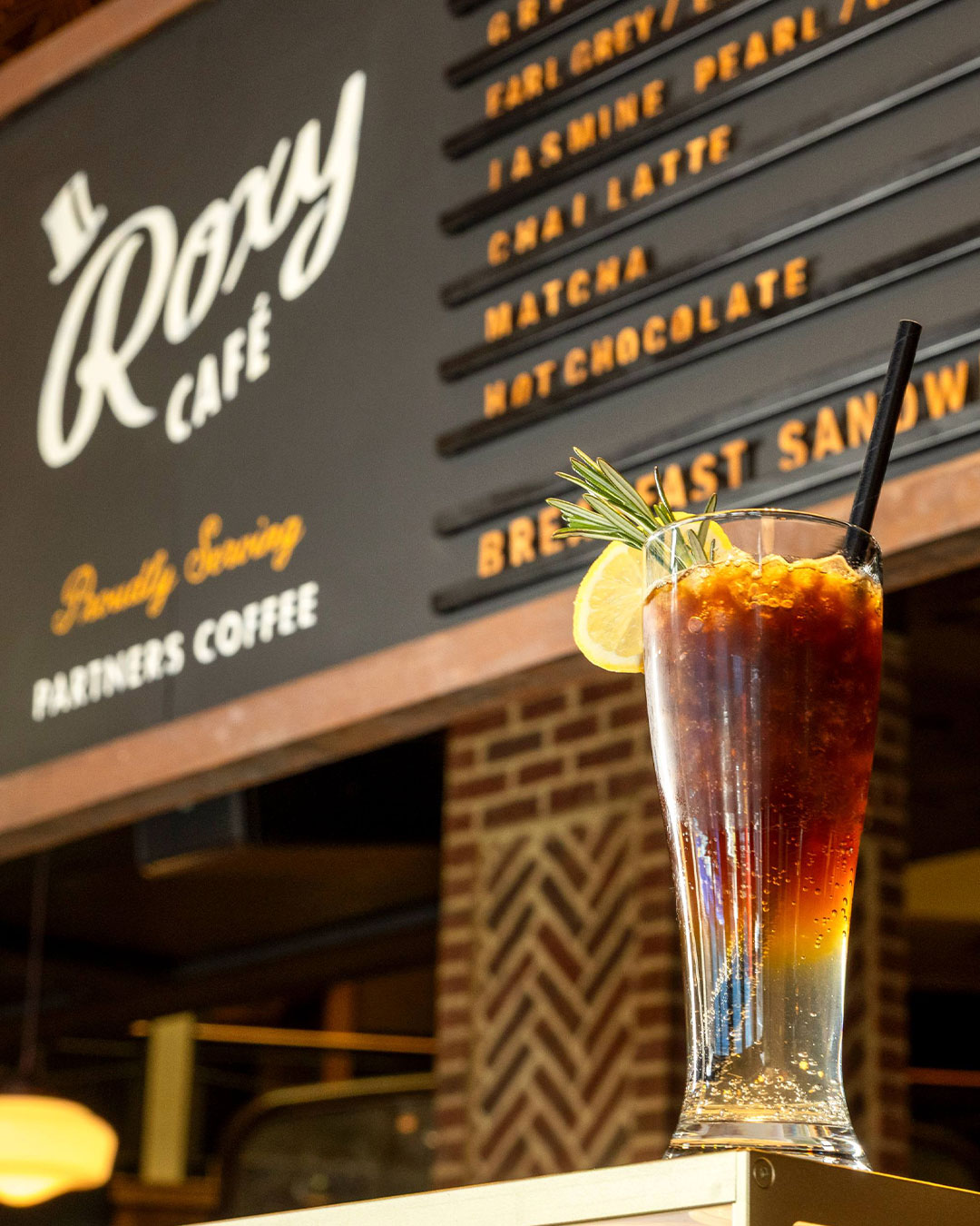 An iced specialty coffee drink garnished with lemon and rosemary in front of the Roxy Café NYC menu board featuring Partners Coffee.