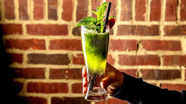 Roxy Café TriBeCa signature green cocktail with crushed ice, mint sprigs, and a straw set against a rustic brick wall background.