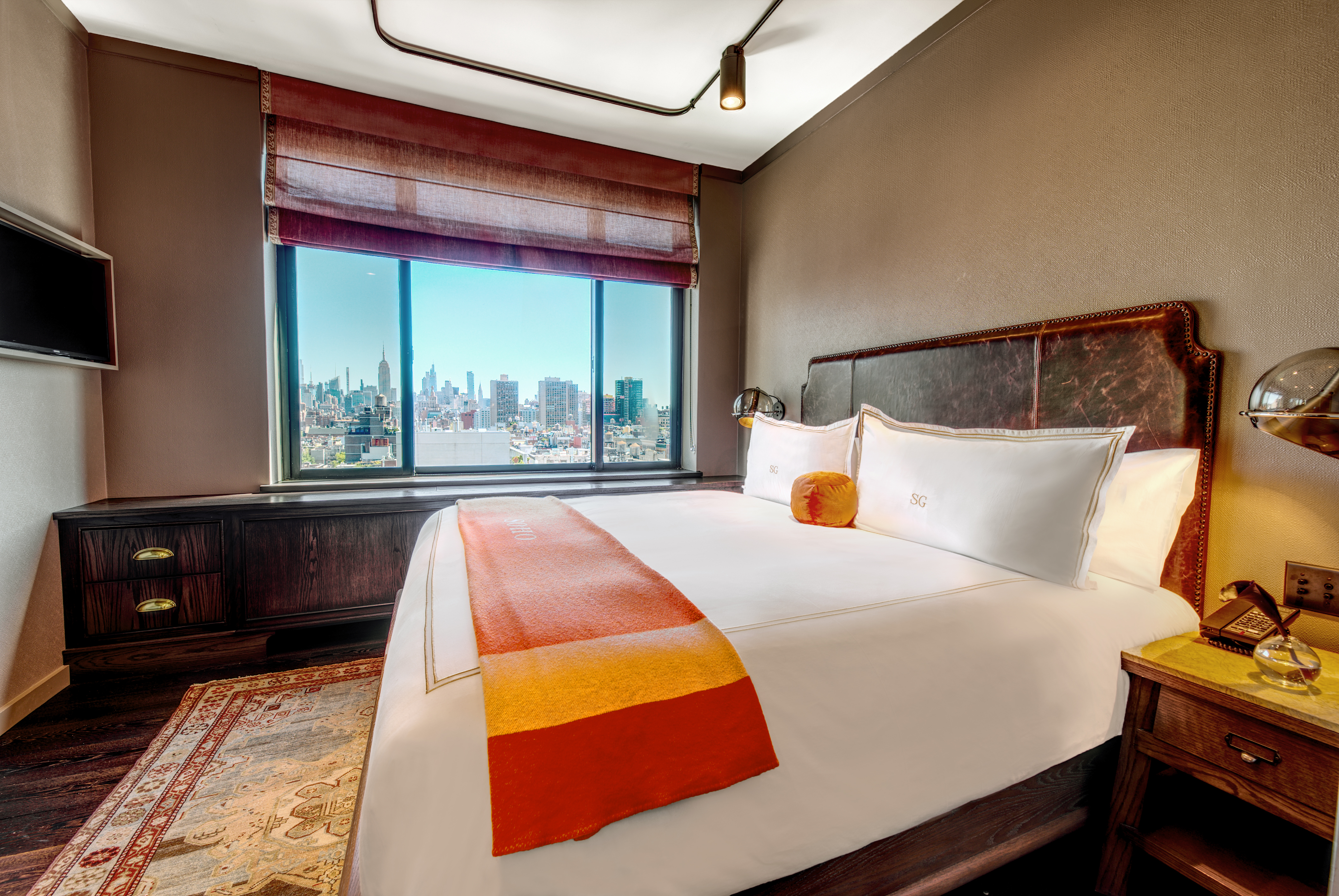 a bedroom in the grand suite. the is a bed, a tv, dresser and a side table. there is also a view of midtown manhattan