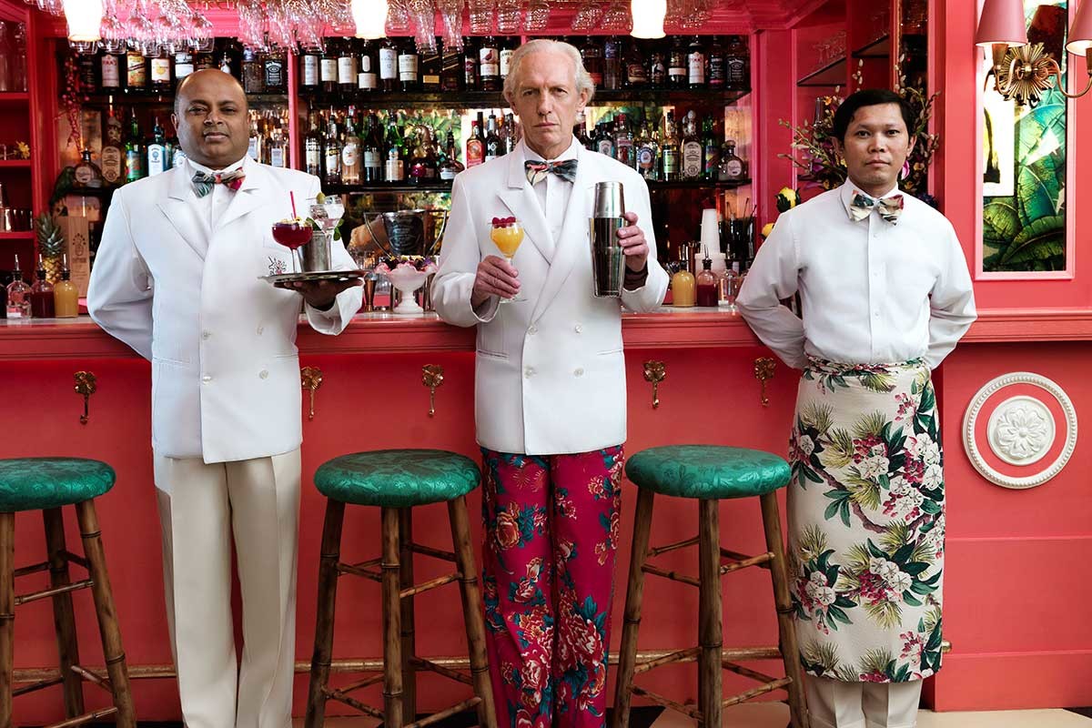 three men in kitchen whites stand in front of a red bar holding different craft cocktails