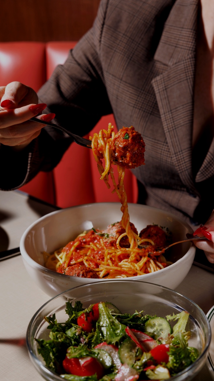 Woman in a brown suit eating spaghetti and meatballs with a salad.