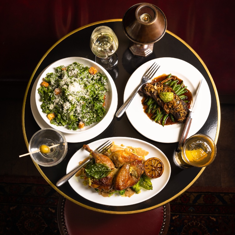 Plates of chicken, steak, and salad on a black table with cocktails at The Django.