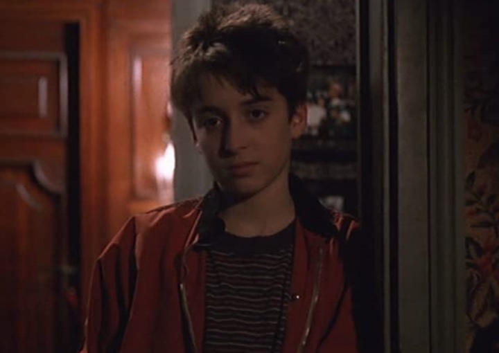 a young boy stands leaning against a door frame with an uninterested look on his face
