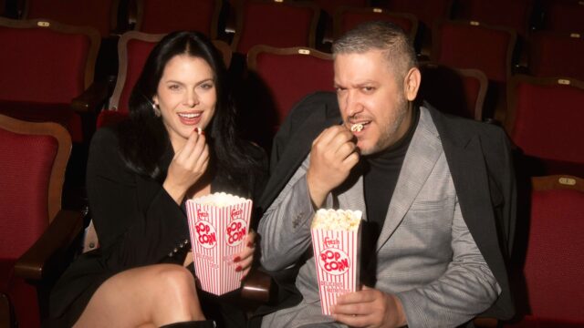 Male and female couple sitting in Roxy Cinema eating popcorn wearing dress clothes.